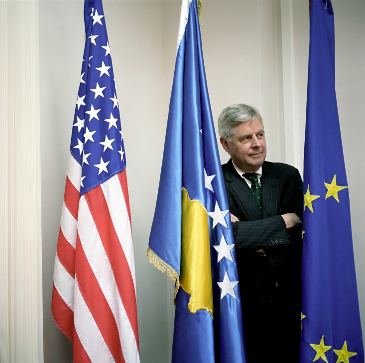 Pieter Feith stands between American, Kosovo, and European Union flags in formal business attire.