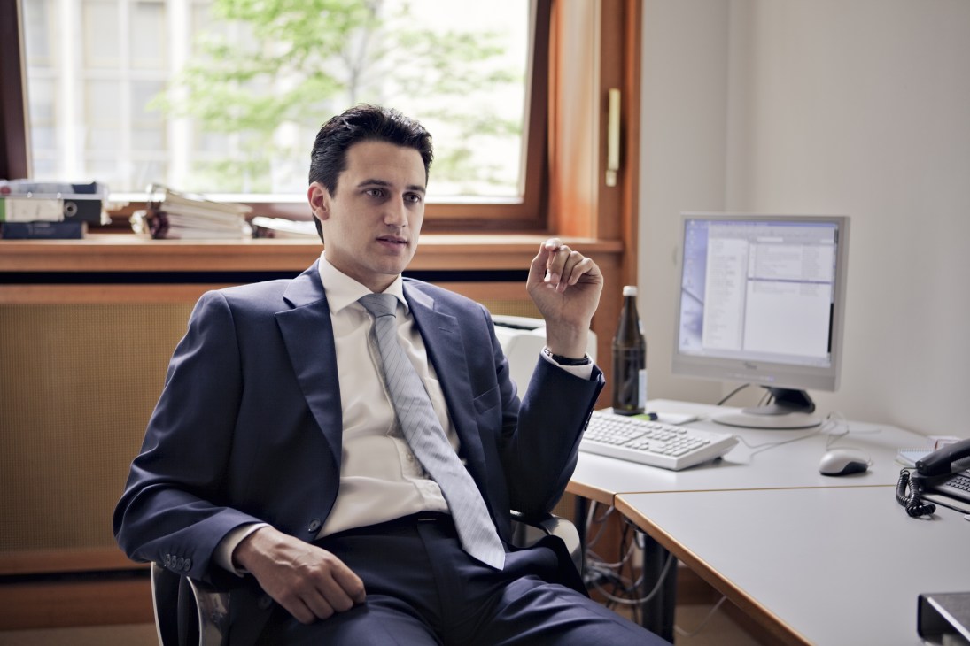 Man in dark business suit and tie sitting at desk in office with computer monitor and window.