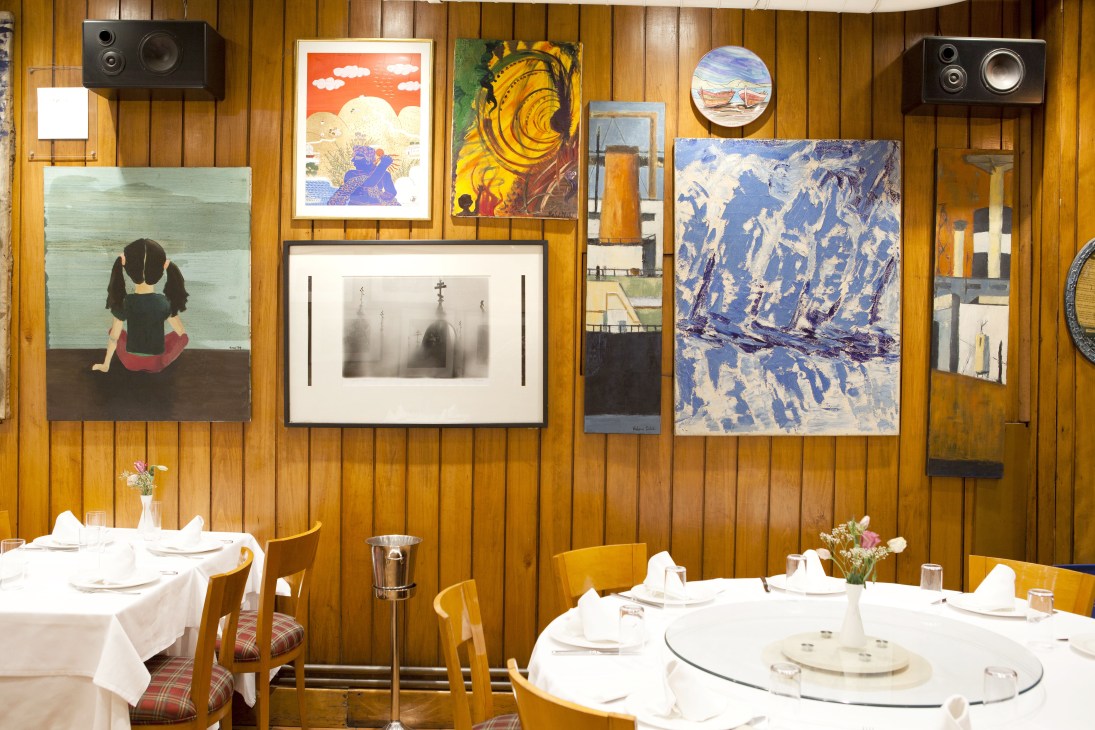 Restaurant interior with wooden walls displaying artworks, set dining tables with white tablecloths and place settings in foreground.