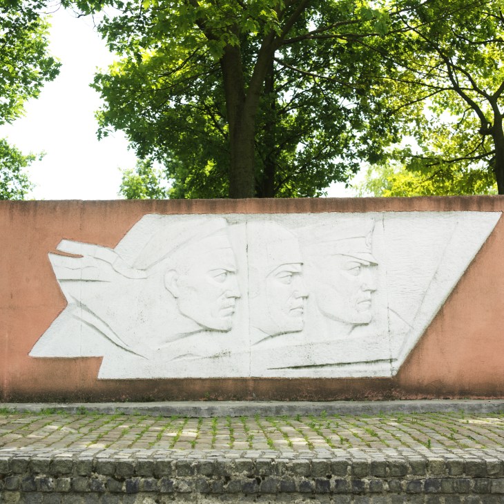 White stone relief sculpture of three sailor profiles carved into a terracotta-colored memorial wall beneath green trees.