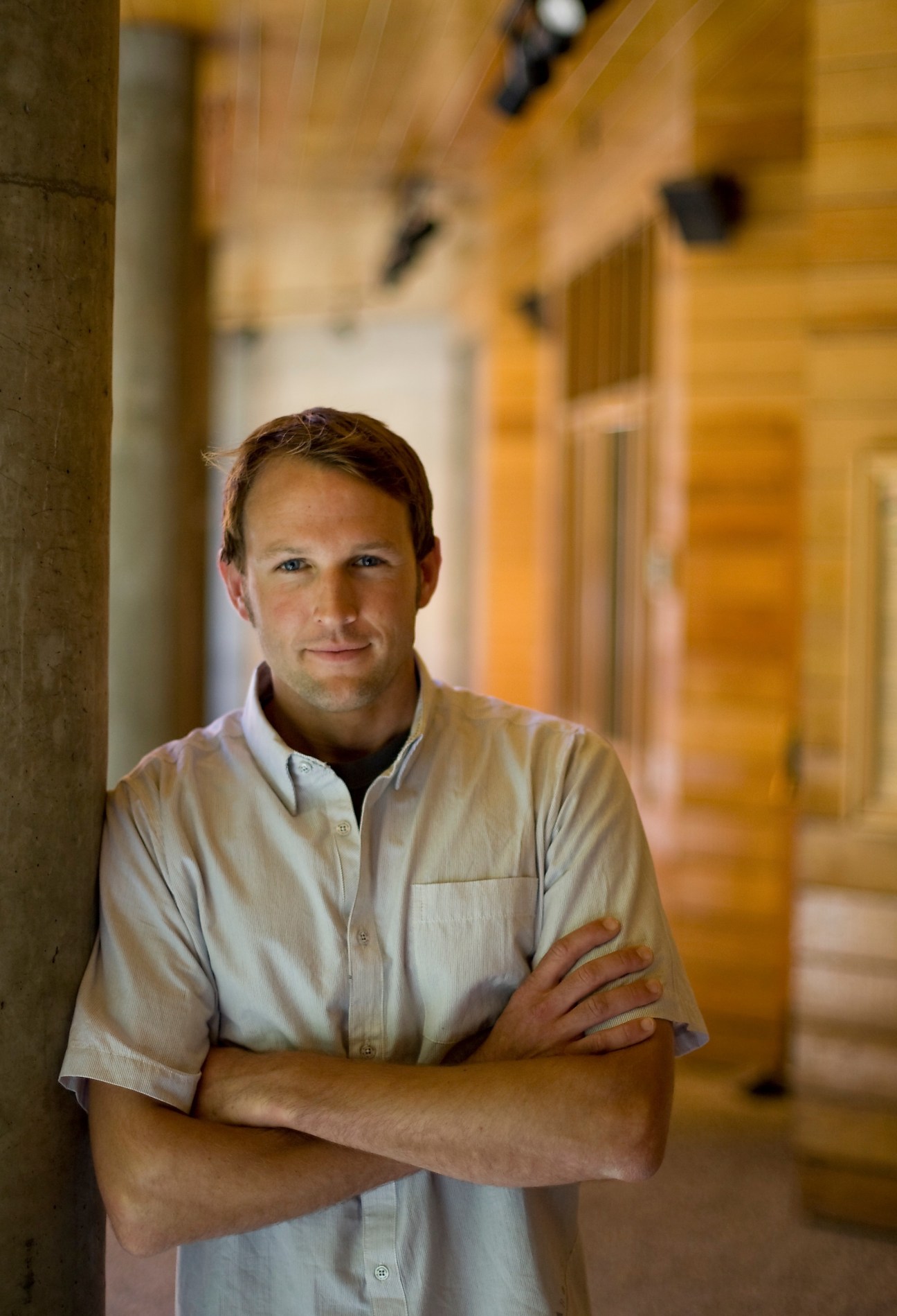 Industrial designer Greg Grenzke stands with arms crossed in a workshop corridor with wooden elements and warm lighting.