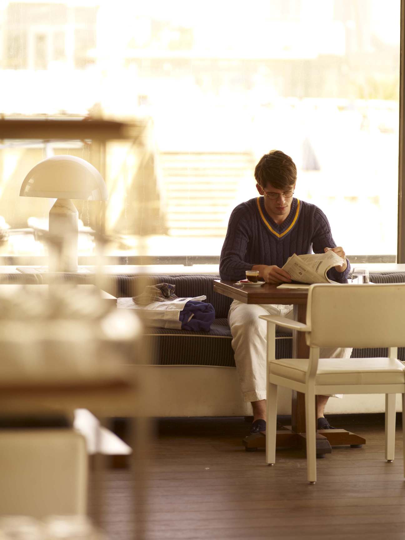 Man wearing burgundy jumper and cream trousers reads magazine indoors in natural light.