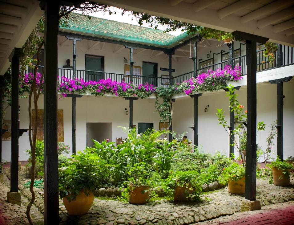 Colonial courtyard with white walls, dark wooden columns, green tile roof, and vibrant magenta bougainvillea flowers.