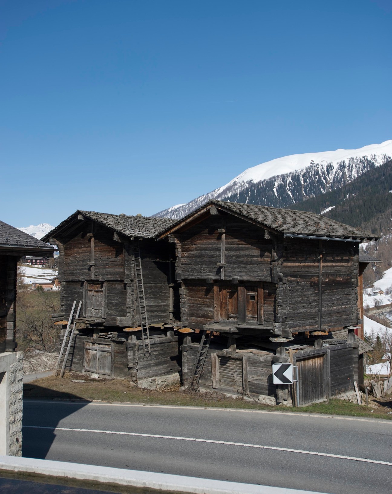 Traditional wooden chalet with dark weathered timber and slate roof set against snow-capped alpine mountain.