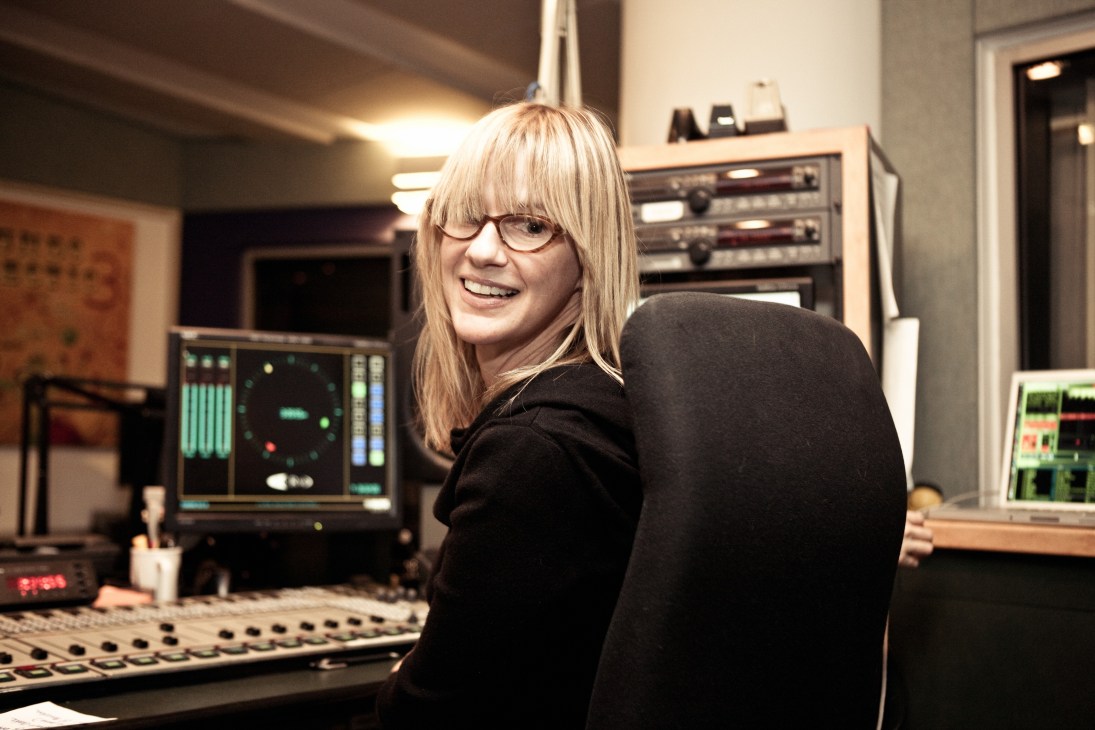 Anne Lit smiles at the camera in a KCRW broadcasting studio with audio equipment and mixing console behind her.
