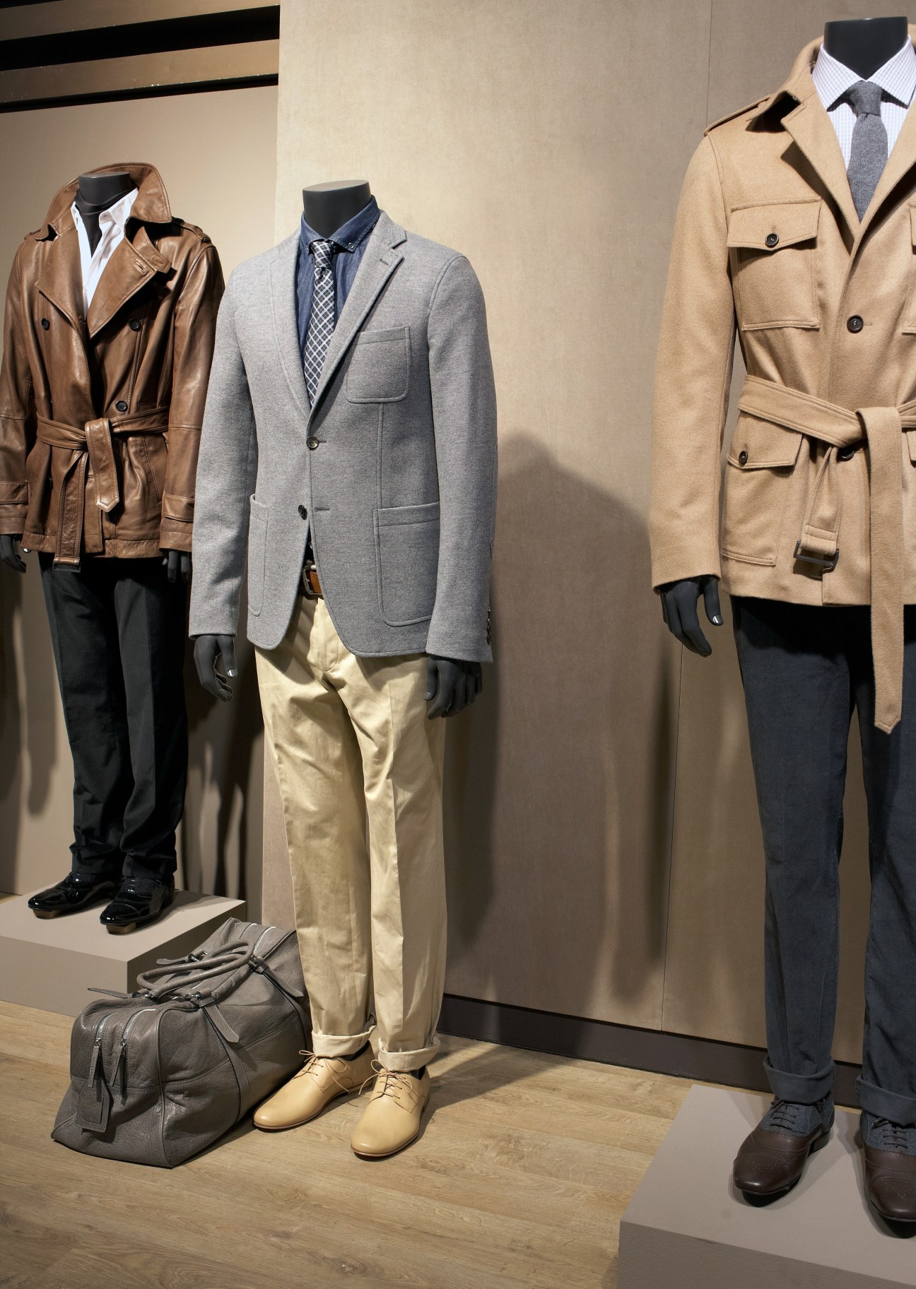 Three mannequins displaying menswear outfits featuring blazers, trousers, and accessories on a wooden floor.