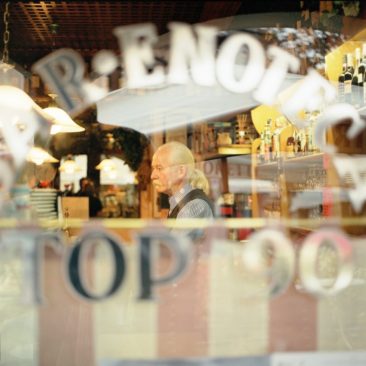 Enzo Marcer, owner of Top 90 bar in Belluno, sitting inside his establishment viewed through a window.