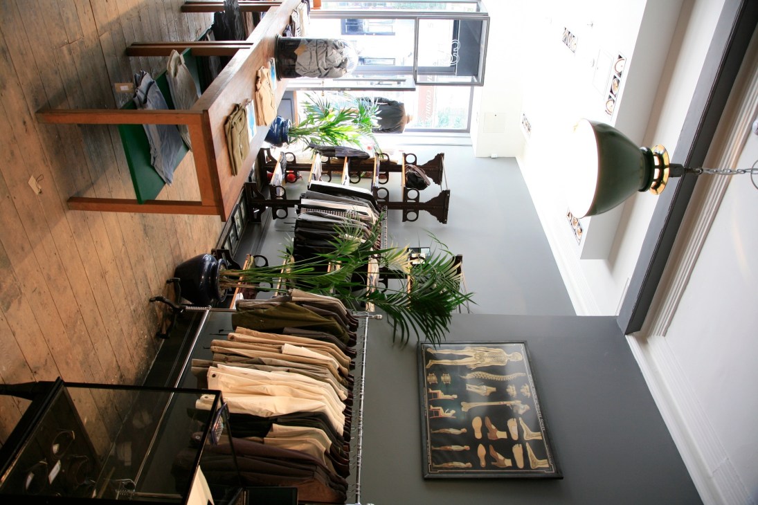 Oliver Spencer menswear store interior with clothing displayed on black racks, plants, anatomical artwork, and natural lighting.