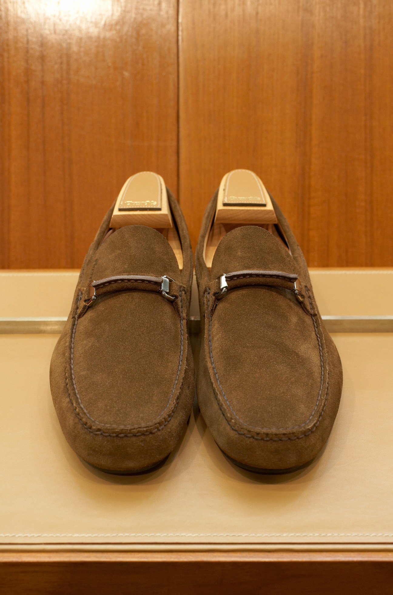 Pair of brown suede loafers with metal bit detail displayed on cream surface against wooden background.
