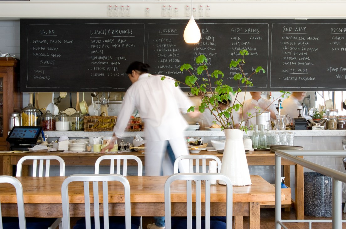 Server in white shirt working at the counter of Allô restaurant with menu board and open kitchen visible behind.