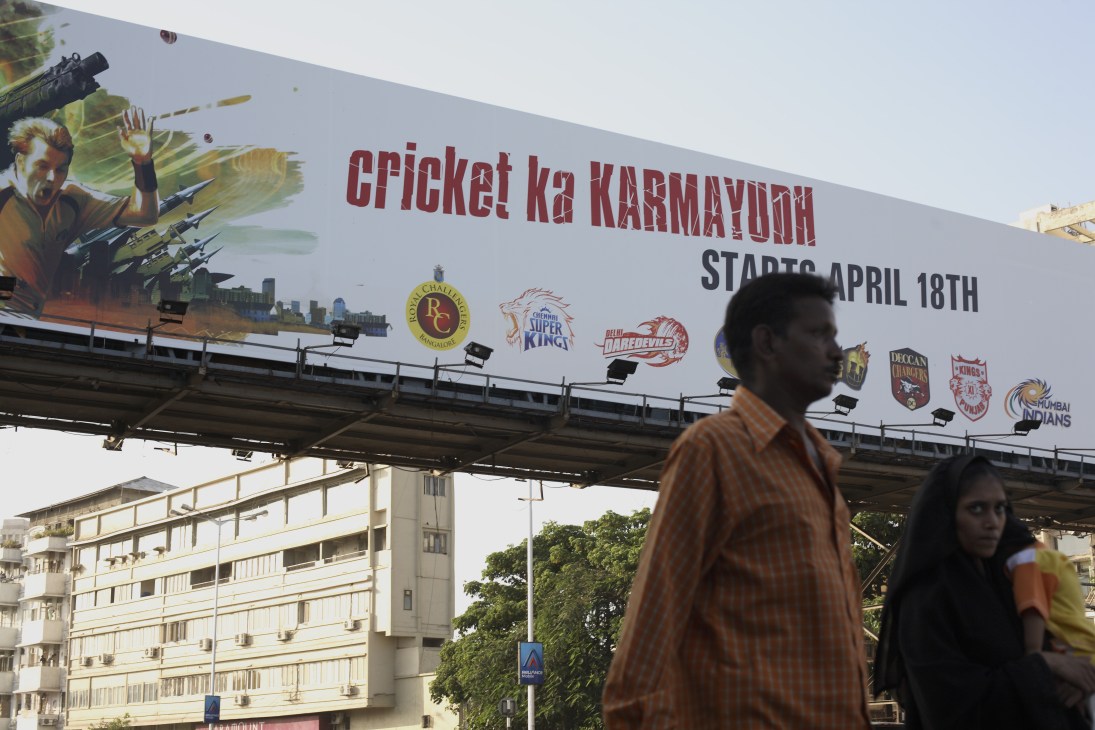A large billboard advertising cricket tournament with military imagery and team logos, with pedestrians walking beneath it on a Mumbai street.
