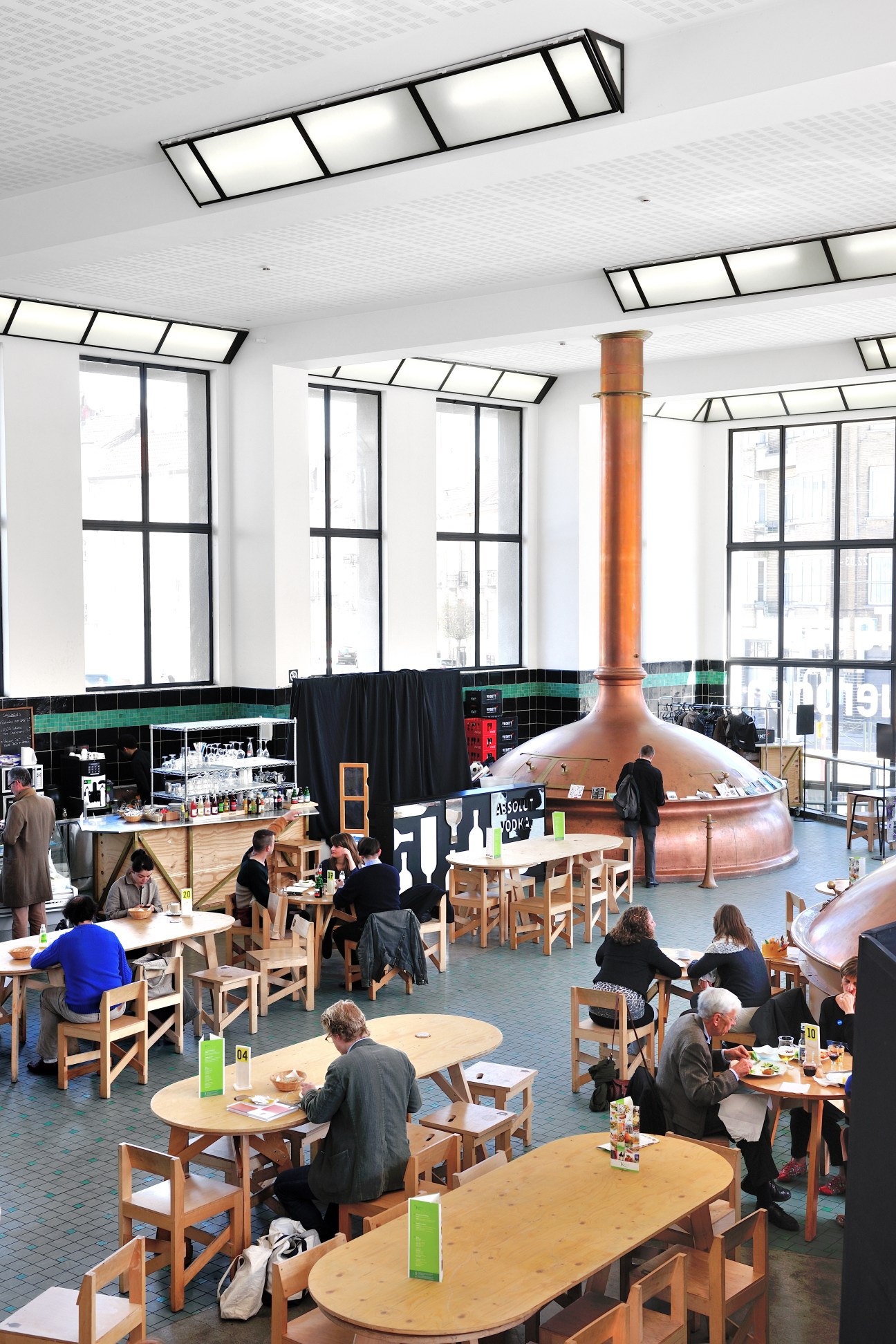 Interior of Wiels museum eatery with copper brewing vessel, skylights, and diners at wooden tables.