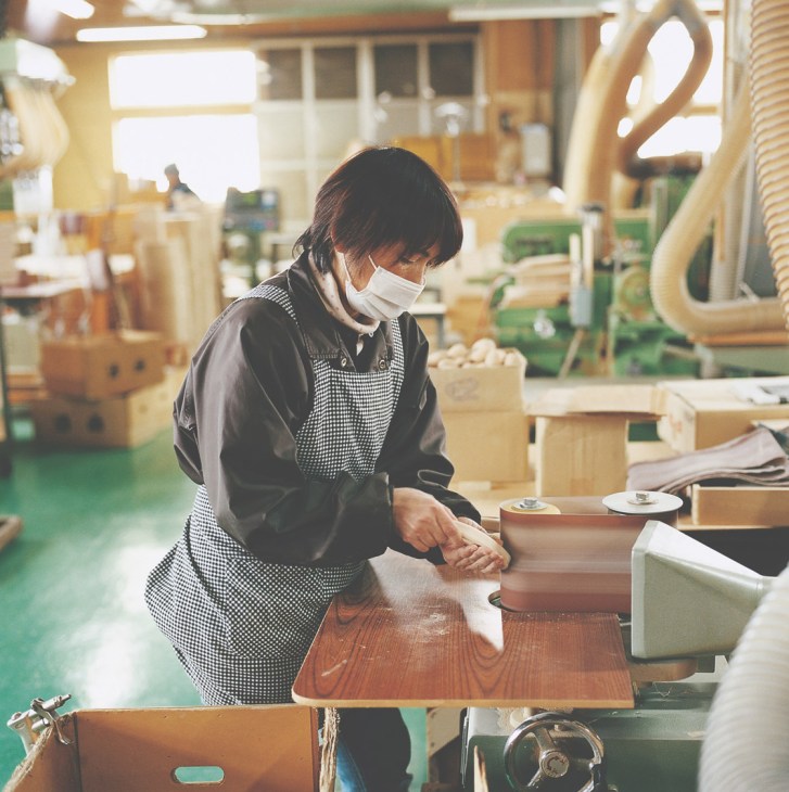 A worker in a checkered apron and face mask polishes a wooden plank in a workshop filled with wooden toy pieces and machinery.