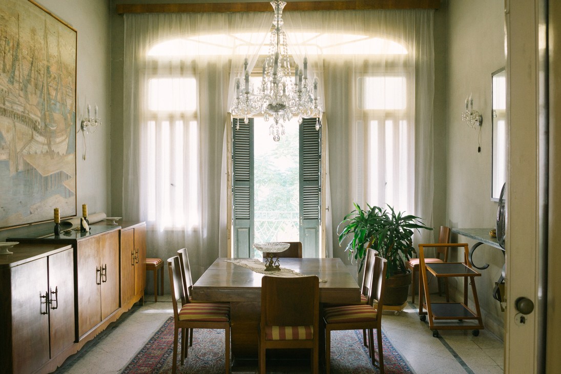 Elegant dining room with crystal chandelier, wooden table, arched windows with green shutters, and ivory curtains.