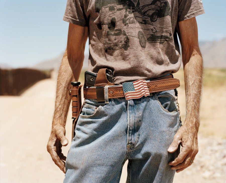 Man wearing a graphic t-shirt and jeans with a brown leather belt featuring an American flag buckle and holstered handgun.