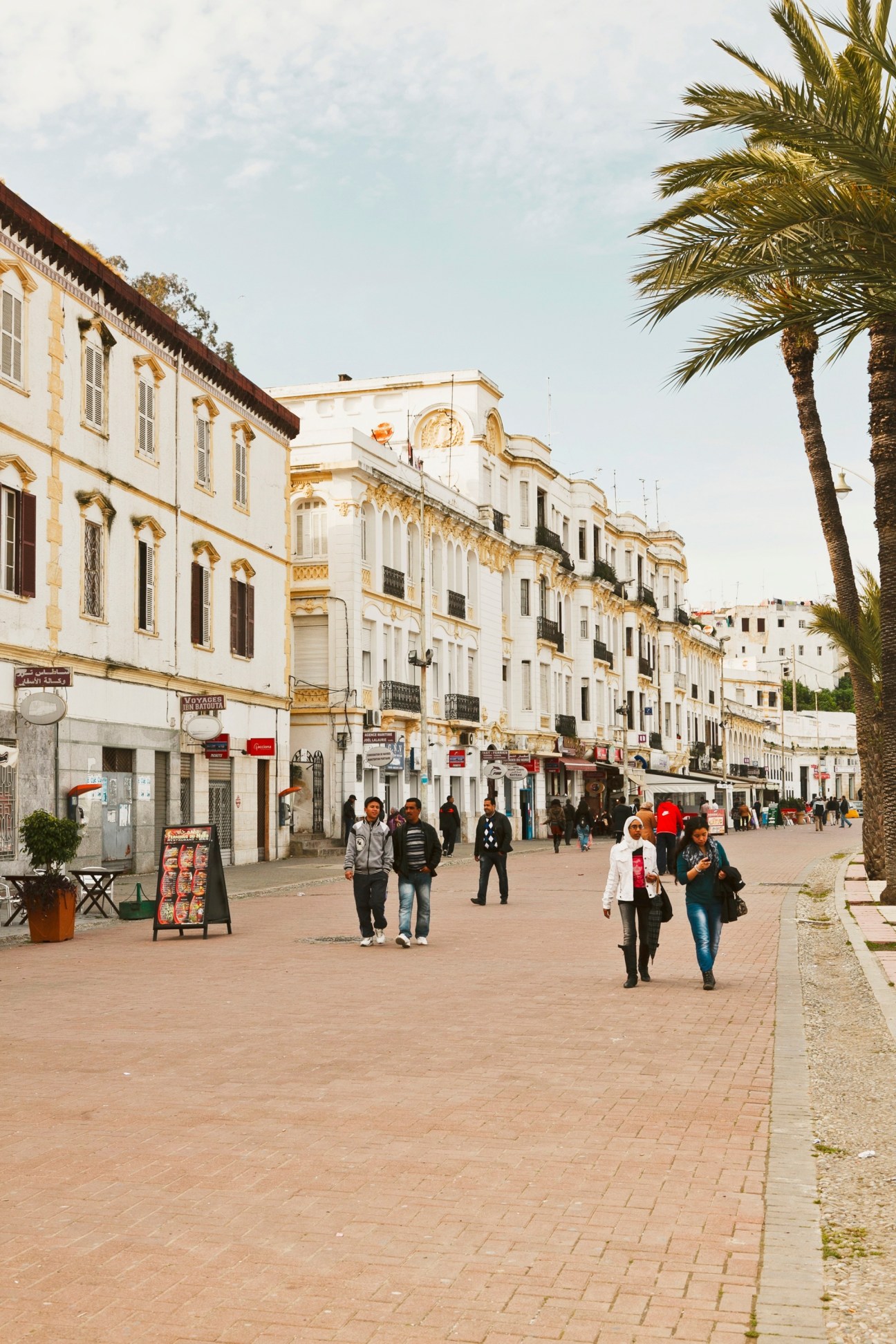 Pedestrians walking along Avenue Mohammed VI in Tangier with colonial-era buildings and a palm tree.