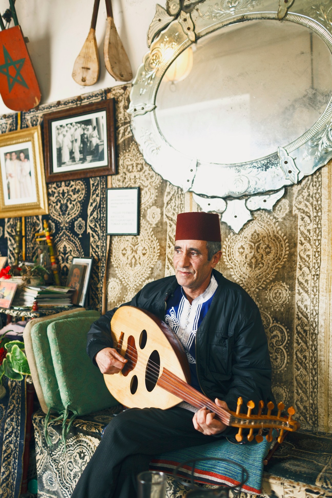 A man wearing a red fez and traditional Moroccan clothing plays an oud in a richly decorated room with patterned wallpaper and ornate mirror.