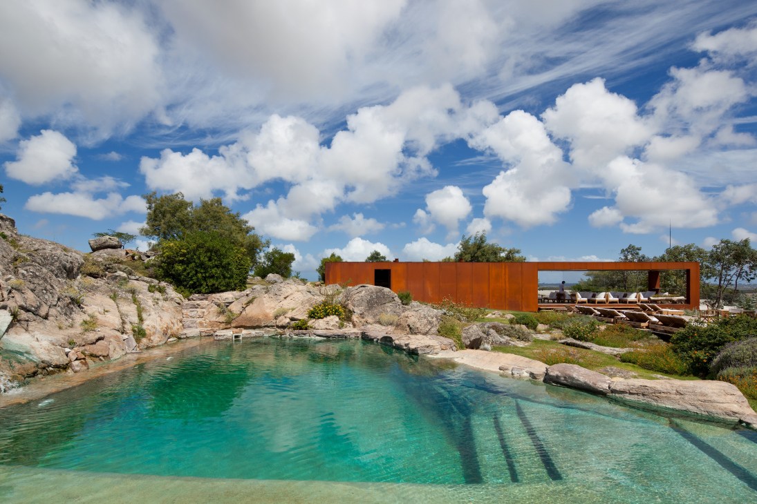 A turquoise pool carved into rocky terrain with a modern rust-colored pavilion structure overlooking it.