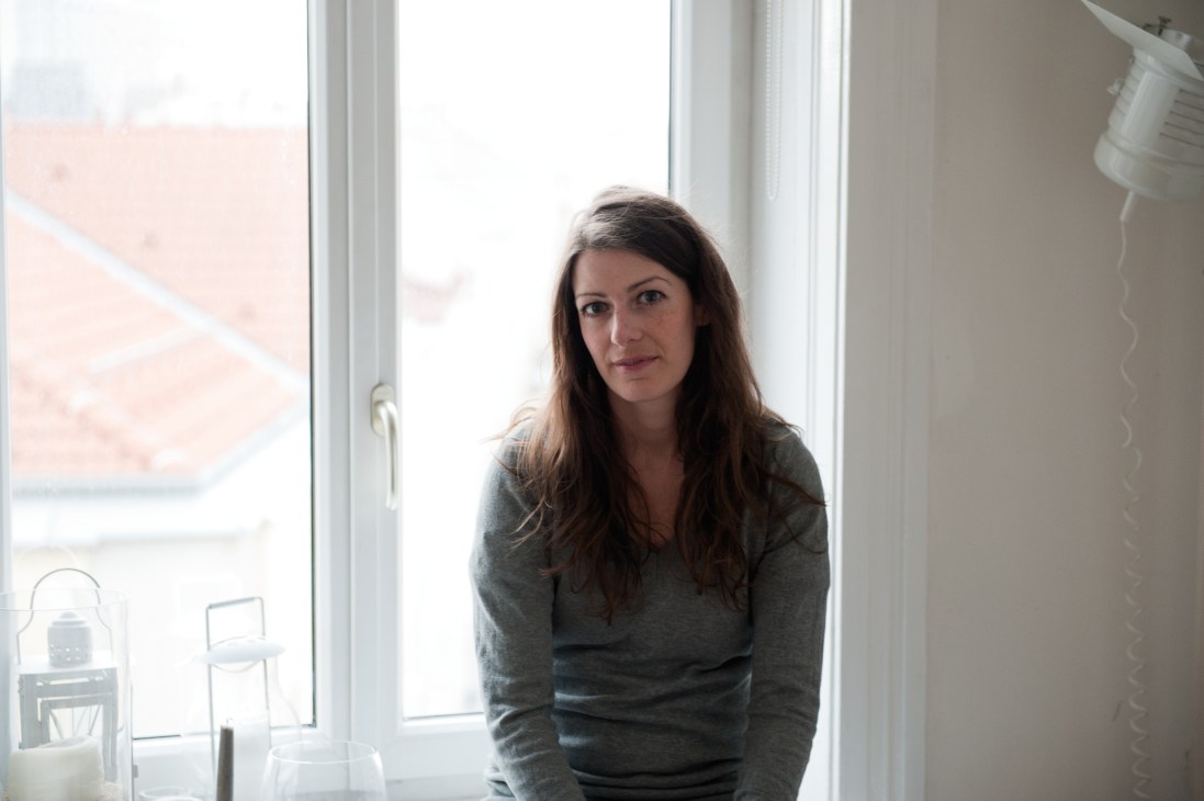 Woman with long dark hair standing by a bright window in a modern apartment interior.