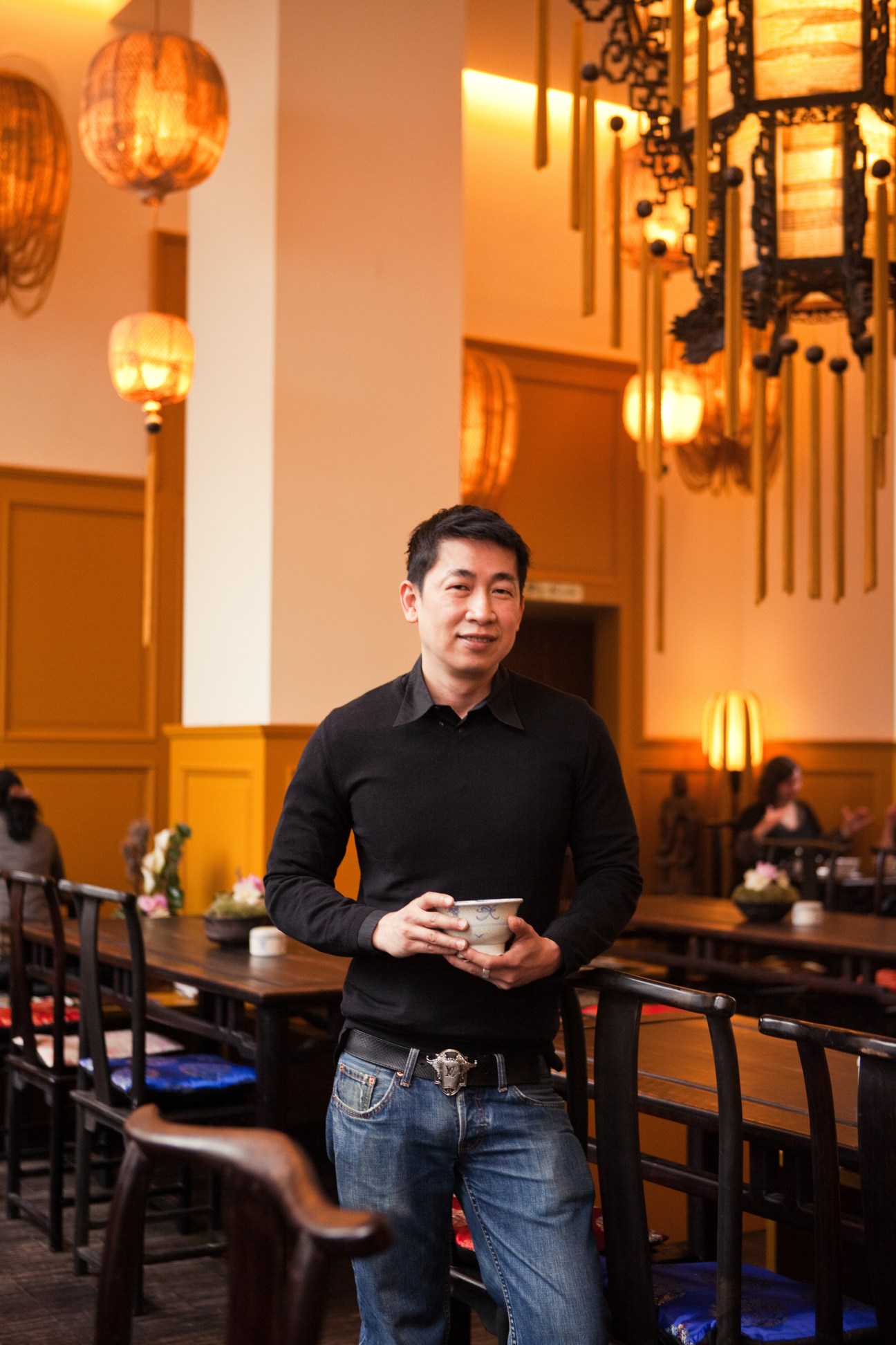 Truong Si An stands in Chen Che tea house holding a small cup, surrounded by traditional Asian lanterns and wooden furniture.