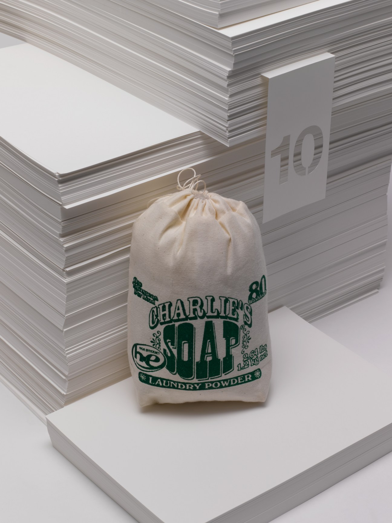 Charlie's Soap laundry powder in a cream linen bag with green printed branding, displayed against stacked magazines.