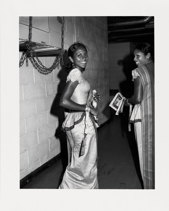 Two smiling Indian women track athletes stand backstage in light-colored athletic wear holding photos.