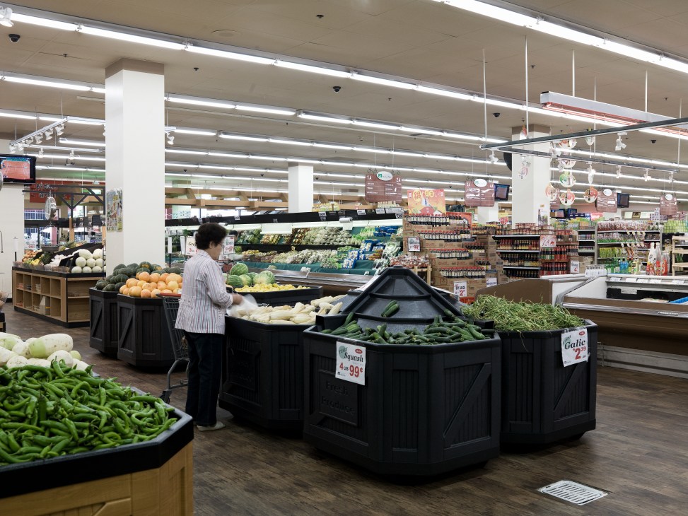 A supermarket produce section with fresh vegetables displayed in black bins and shelving, with a staff member arranging items.