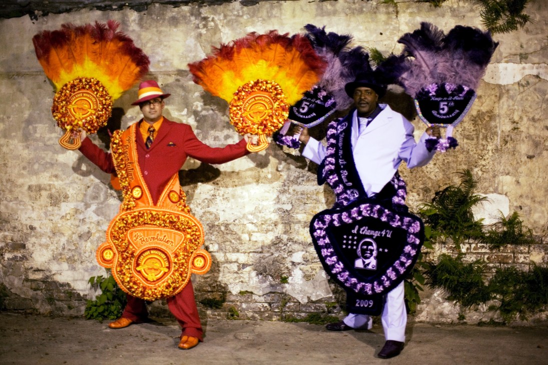 Two men in elaborate colorful carnival costumes with large feathered headdresses pose against a weathered brick wall.