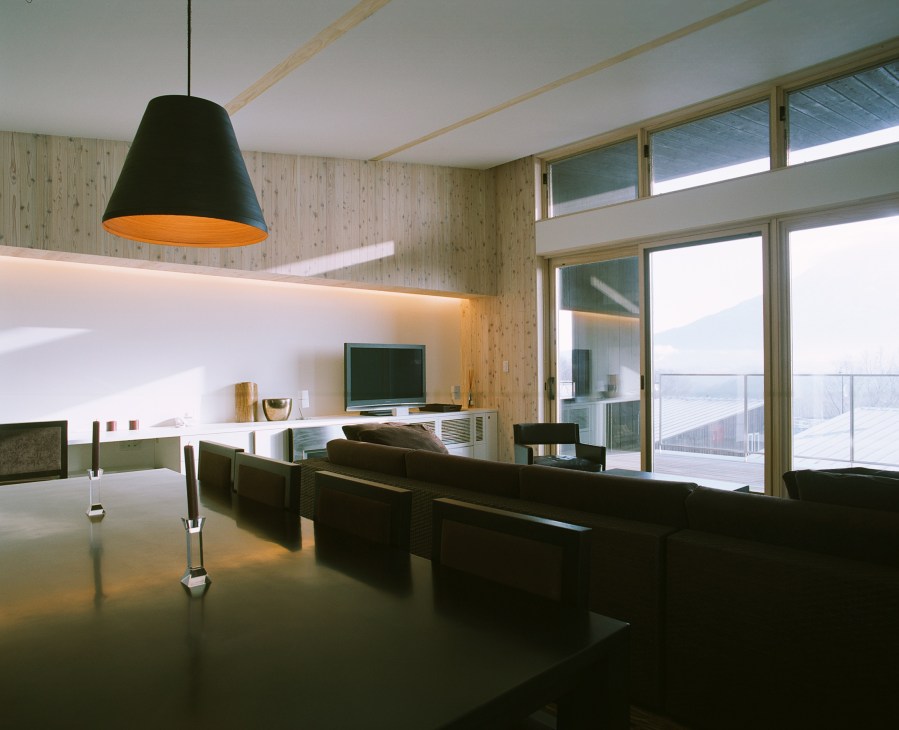 Modern living room with wood-paneled walls, black pendant light, dark furniture, and floor-to-ceiling windows overlooking mountain landscape.
