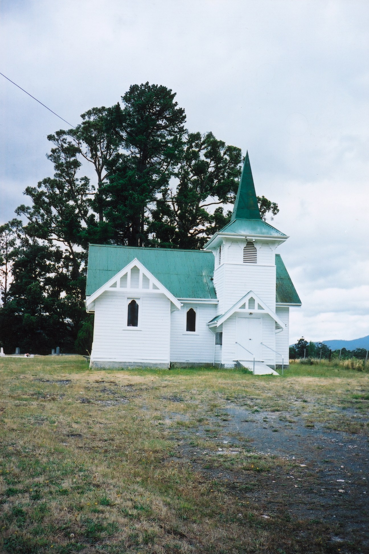 White wooden church with green roof and steeple in Glen Huon, Tasmania, with large tree behind.