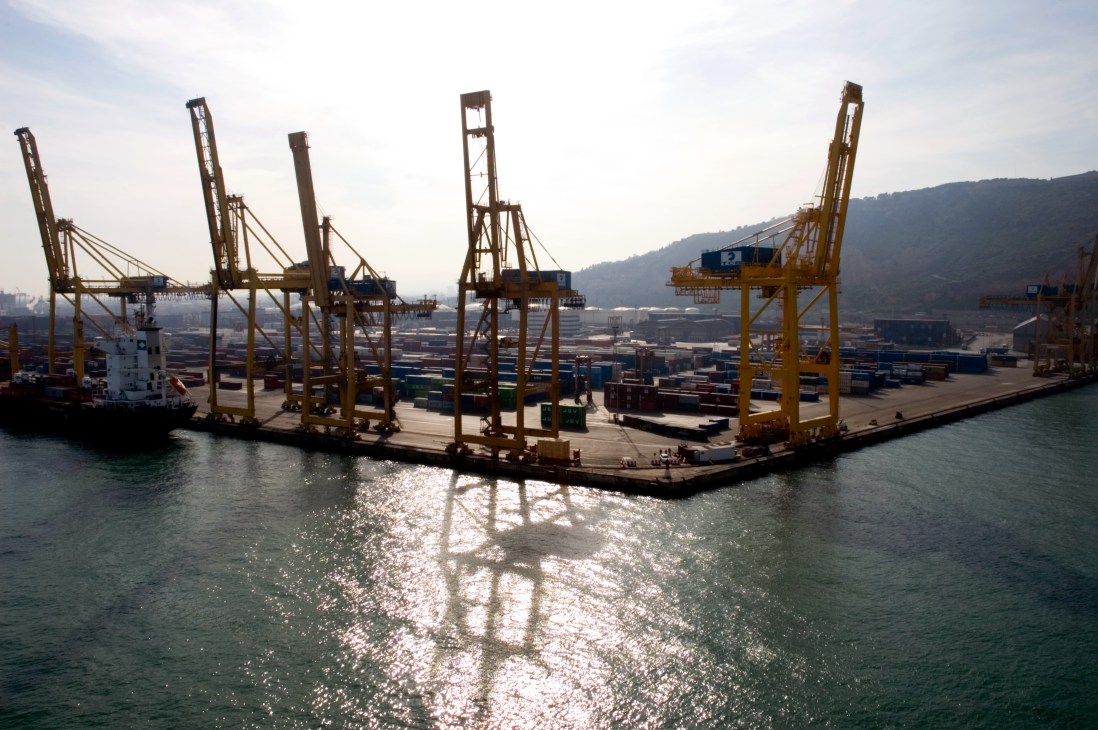 Container cranes and stacked cargo at Barcelona port viewed from the water.