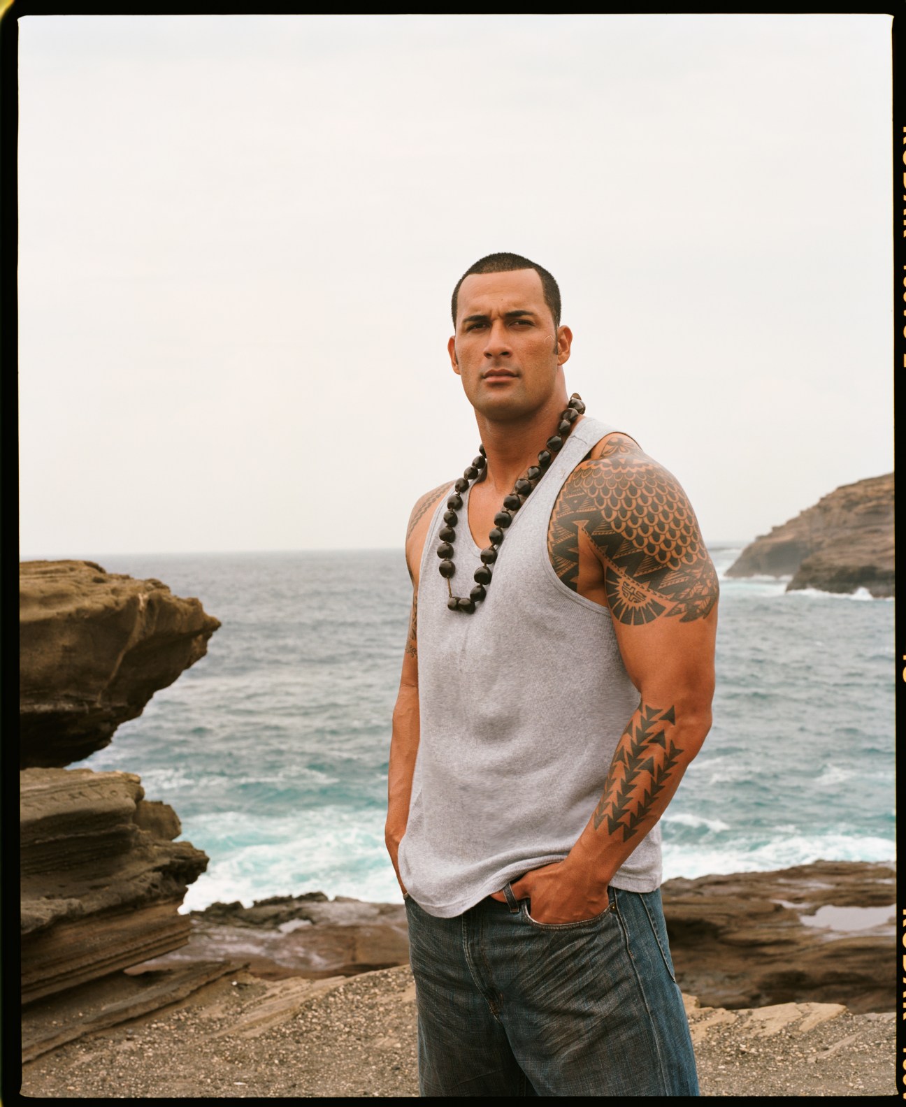 Local businessman Kaui Kauhi standing on rocky Hawaiian coastline wearing traditional lei and tank top with tribal tattoos.