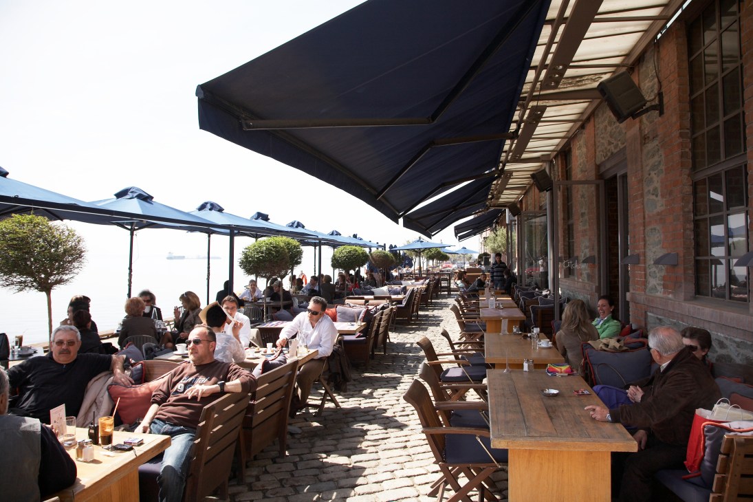 Outdoor waterfront dining at Kitchen Bar with diners seated at tables under black awnings along Thessaloniki's port.