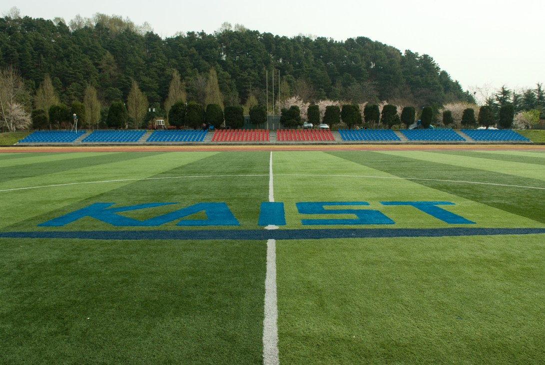 KAIST's sports field with blue and red stadium seating and the university logo painted on the grass.