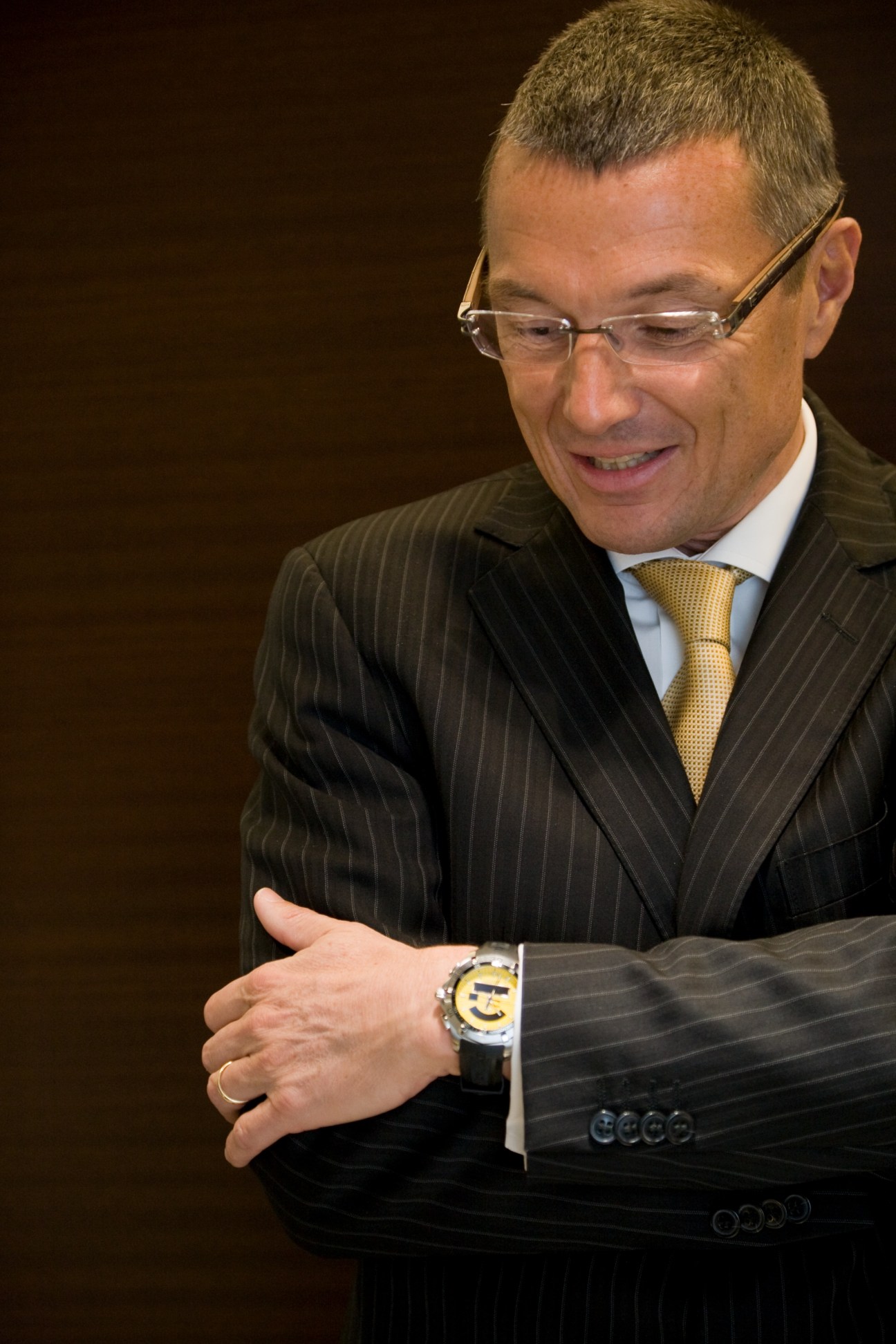 Jean-Christophe Babin, president of TAG Heuer, wearing glasses and a pinstriped suit with gold tie, displays a watch on his wrist.