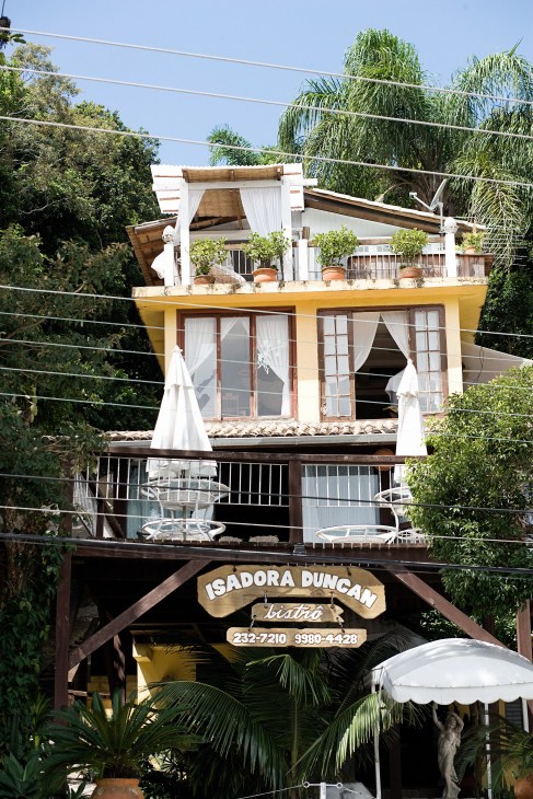 Isadora Duncan restaurant, a yellow and white two-story building with white curtains and potted plants on its rooftop terrace in Barra de Lagoa.