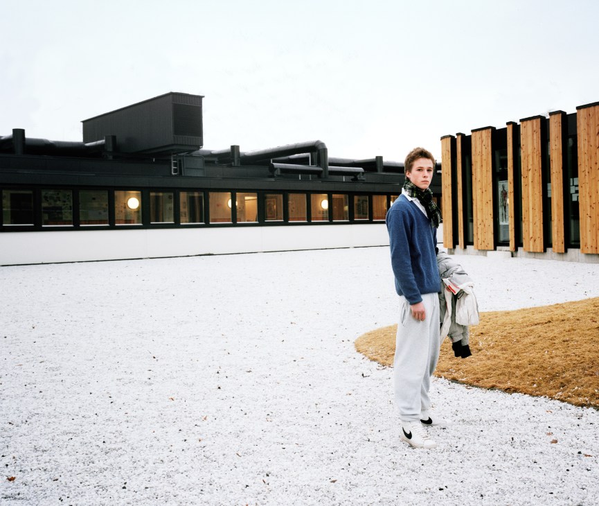 An 18-year-old student stands in the quiet garden area of Oslo International School, with white gravel ground and wooden stools visible.