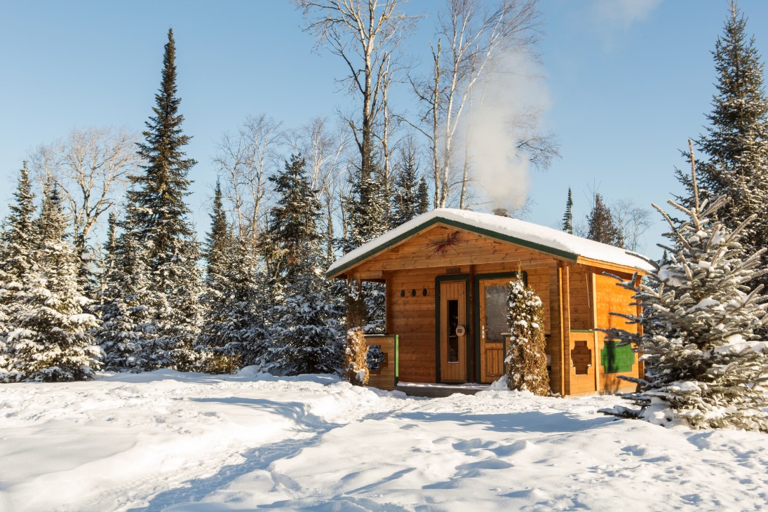 Wooden sauna building with snow-covered ground surrounded by coniferous and birch trees with steam rising from chimney.