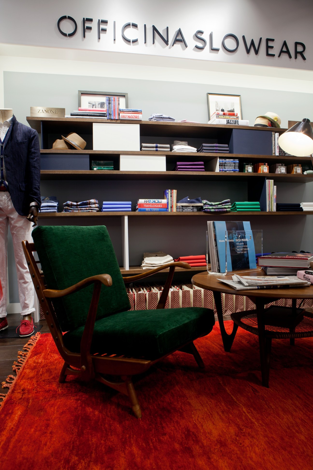 Officina Slowear store interior with shelving displaying folded clothing and accessories, featuring a green mid-century chair on a red rug.