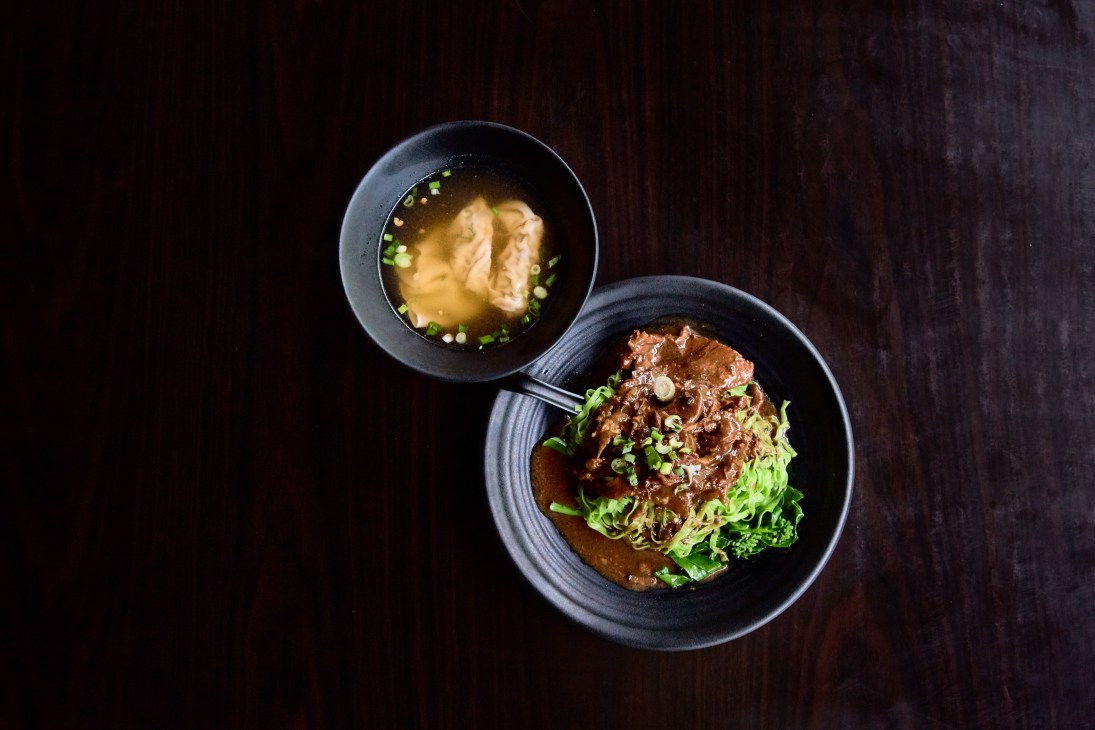 Overhead view of two dark bowls containing spinach noodles with braised beef brisket and garnish on a dark wooden surface.