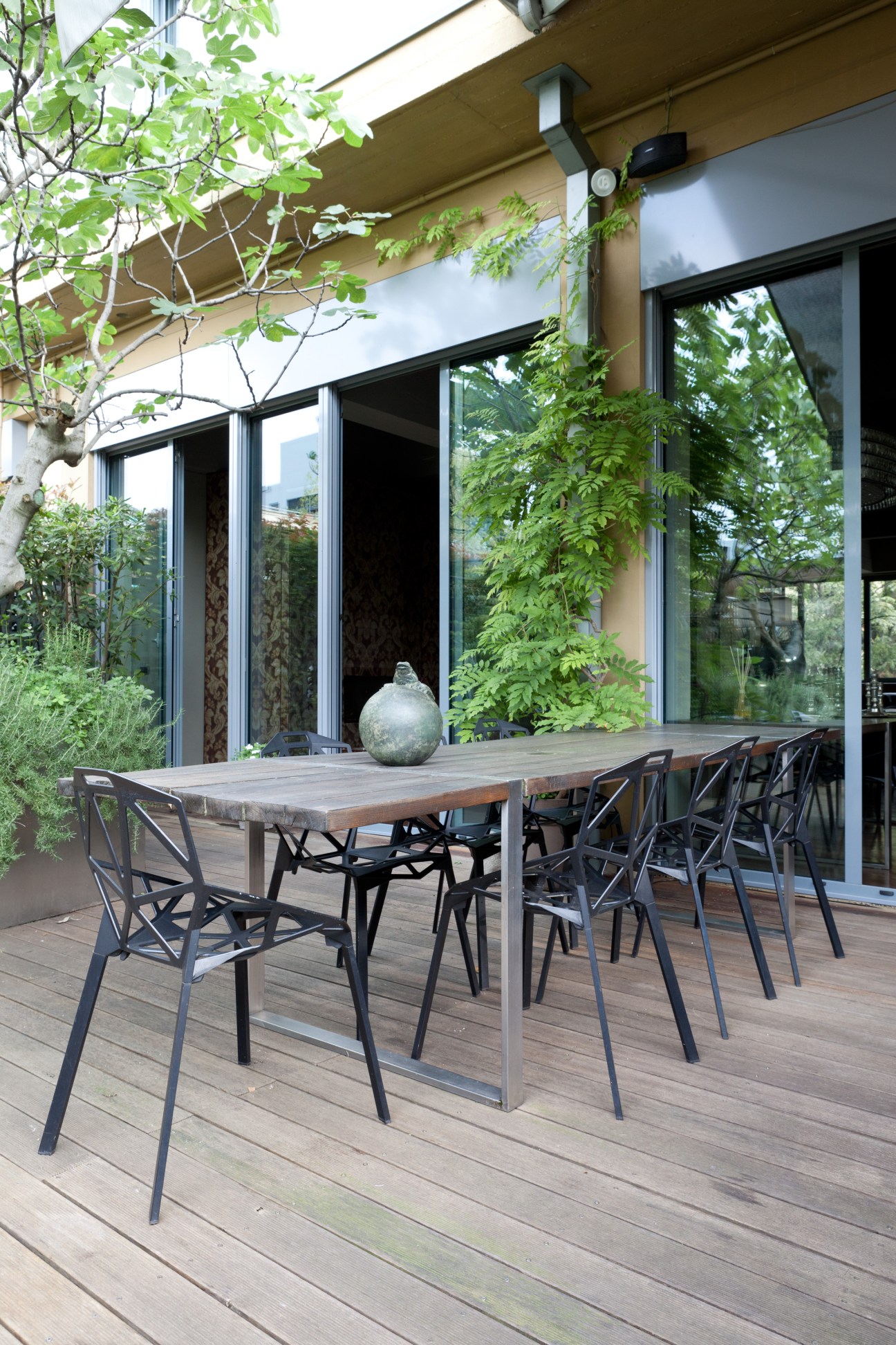 Modern terrace with wooden dining table, black chairs, and large glass doors opening to interior space surrounded by green plants.