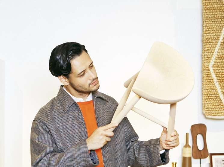Man in Auralee jacket and John Smedley jumper examining a cream-colored chair in a minimalist interior.