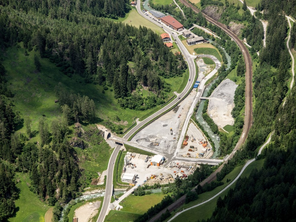 Aerial view of Brenner Rail Tunnel construction site in Alpine valley with forested slopes.