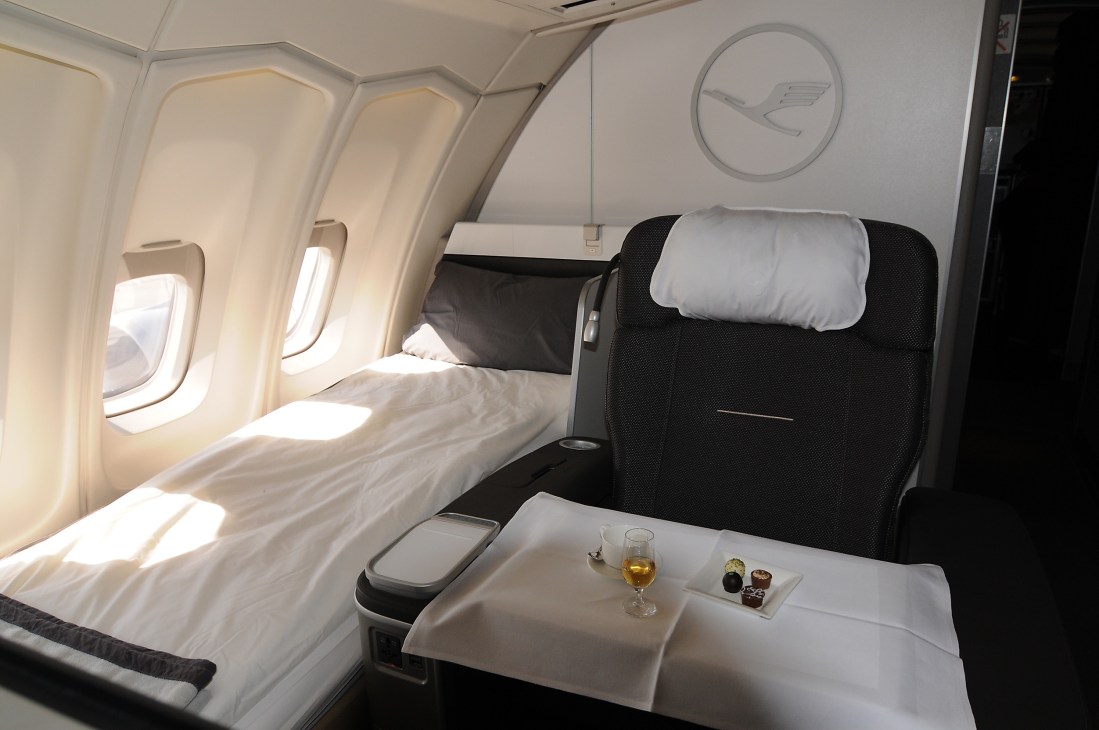 Lufthansa first-class cabin with lie-flat bed, white pillow, black headrest, window seat, and service table with drink and chocolates.