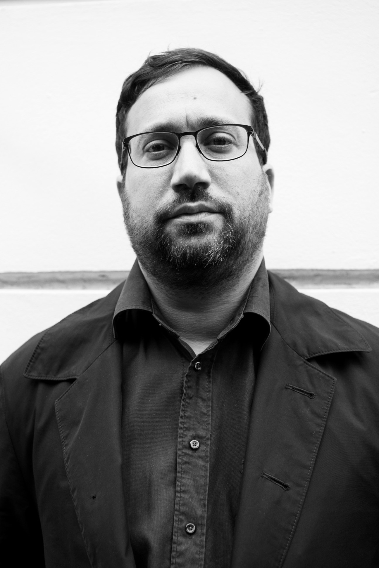 Black and white portrait of a bearded man wearing glasses and a dark shirt against a light background.