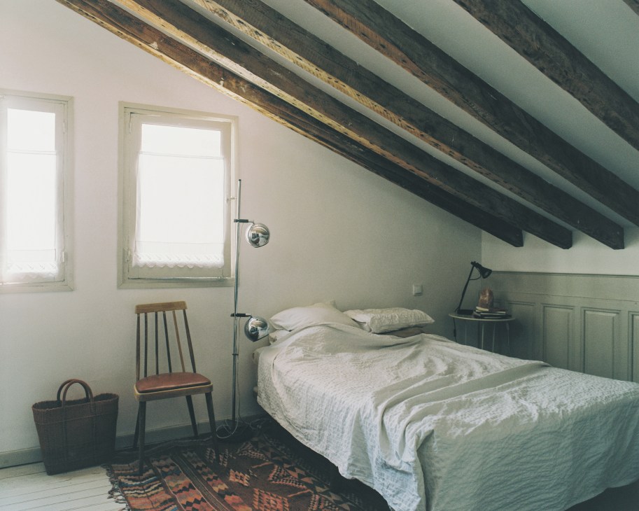 A minimalist bedroom with exposed wooden beams, white walls, green wainscoting, and a simple bed with white linens.