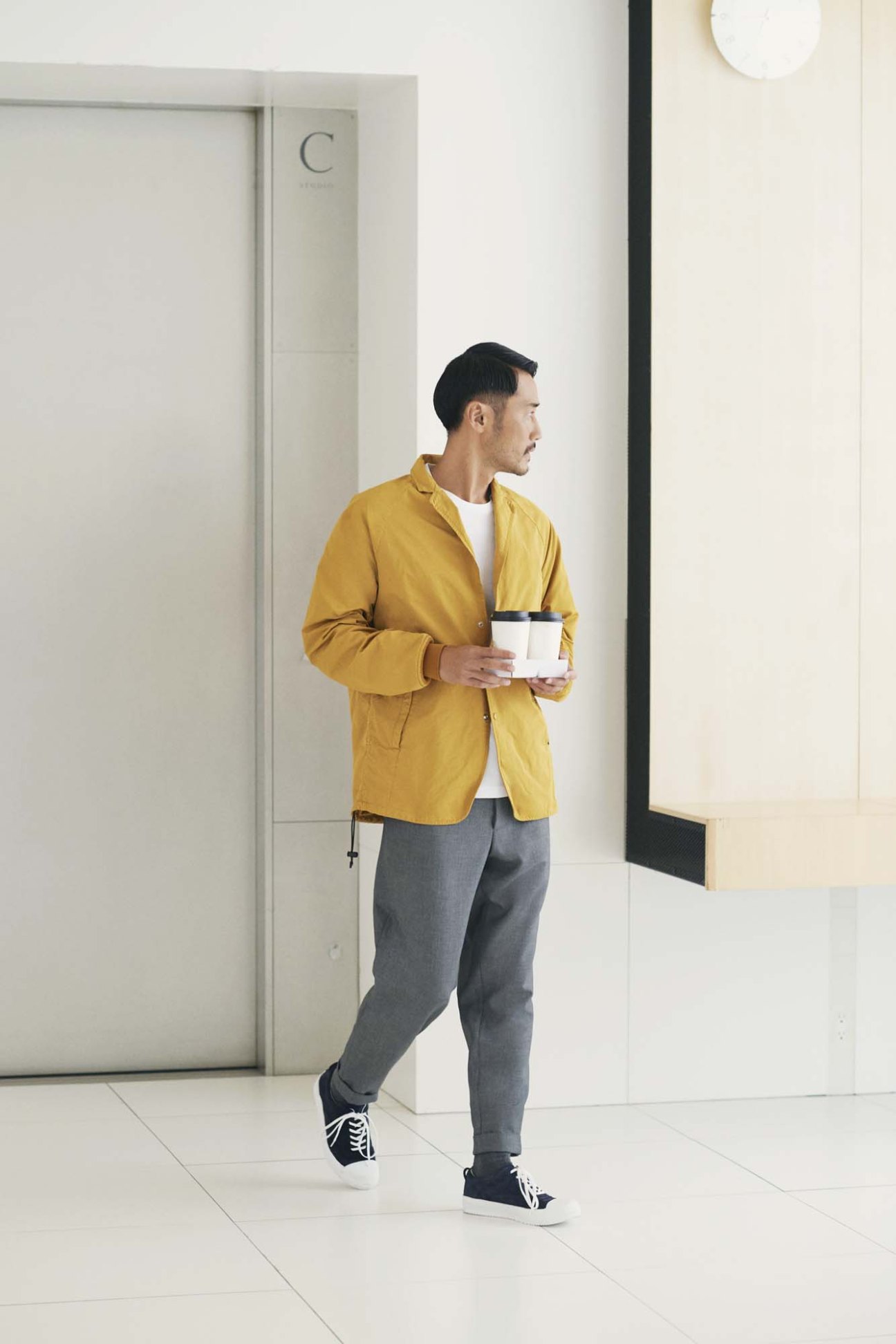 Man in yellow jacket, white t-shirt, grey trousers and black sneakers holding coffee cup in office corridor.