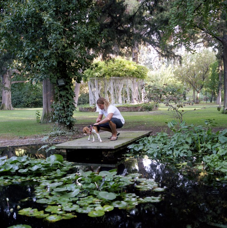 Mark Giraud crouches on a wooden bridge over a pond with his dog in a lush garden.