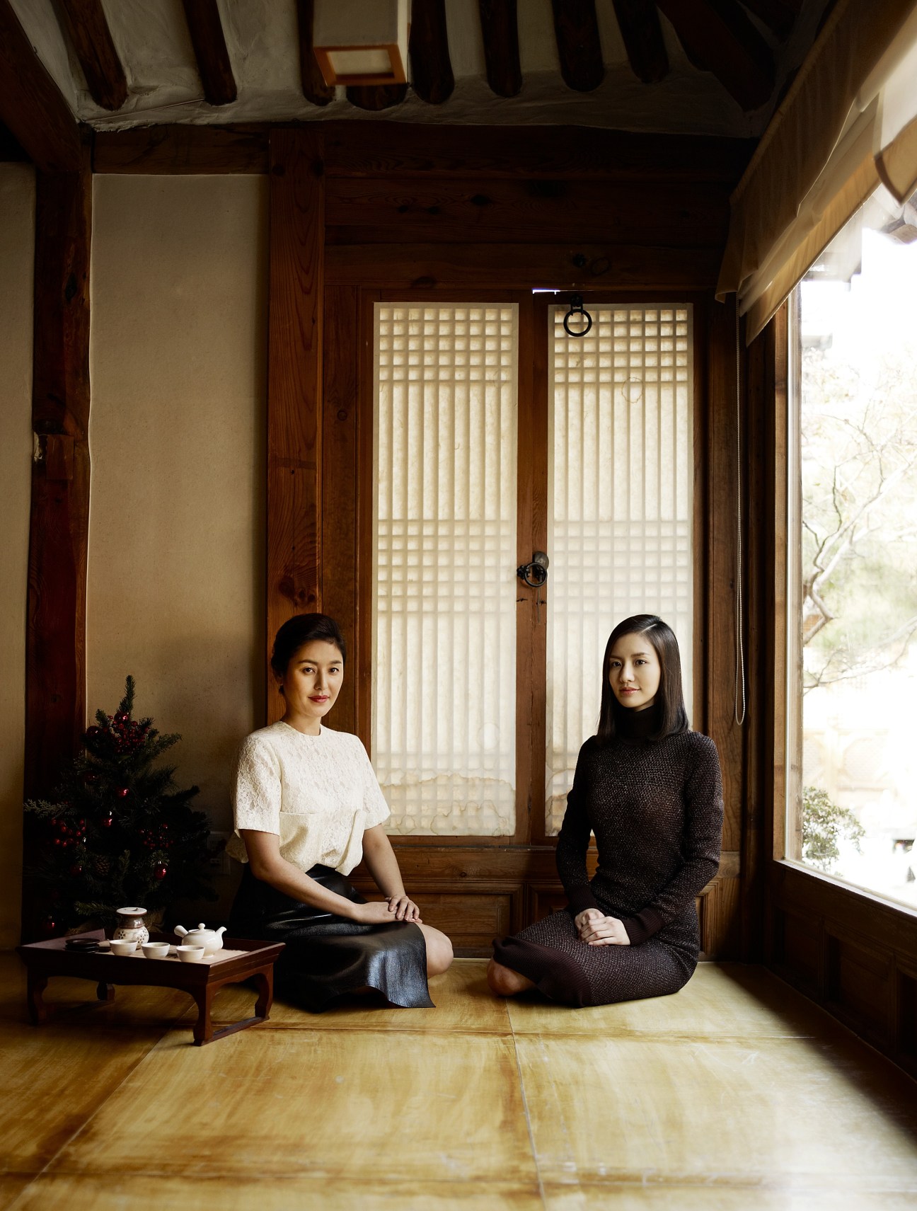 Two women sitting on a wooden floor in a traditional Korean room with latticed doors, one wearing a cream top and the other a black dress.