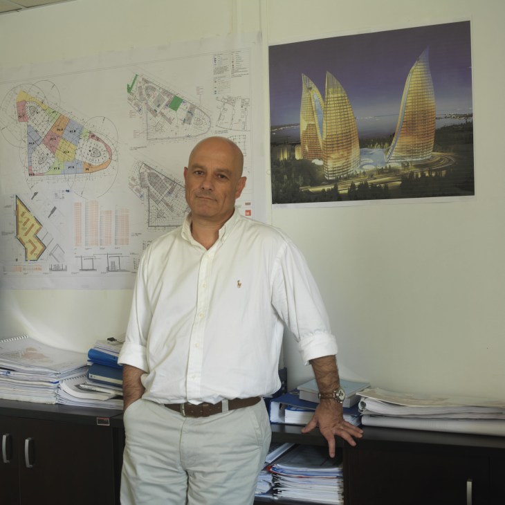 Flame Towers project manager Gokhan Girgin standing in an office with architectural plans and renderings of sail-shaped skyscrapers behind him.